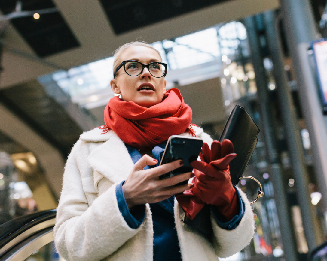 A woman wearing glasses and a red scarf gazes at her phone, engrossed in its contents.
