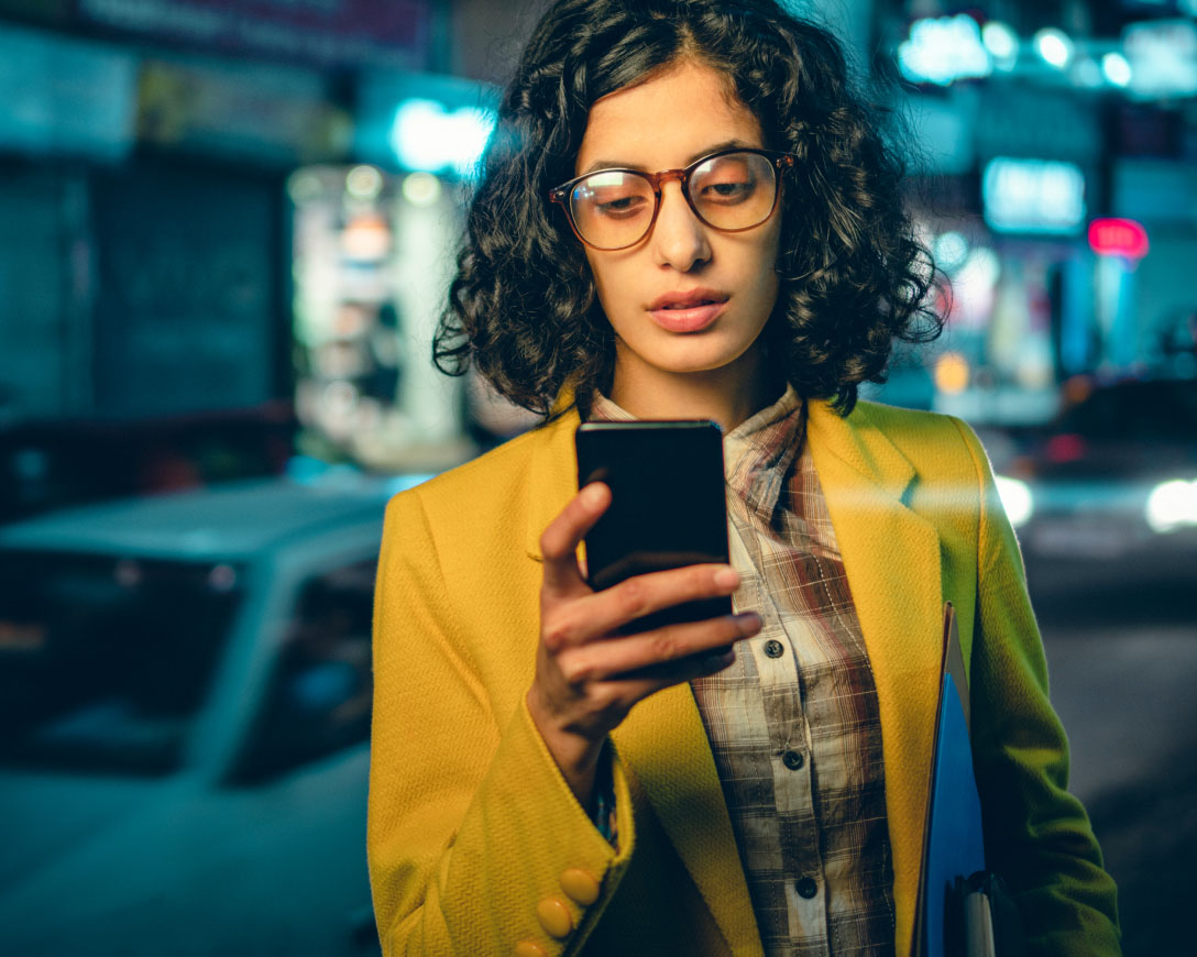 A woman in glasses and a yellow jacket engrossed in her phone, unaware of her surroundings.