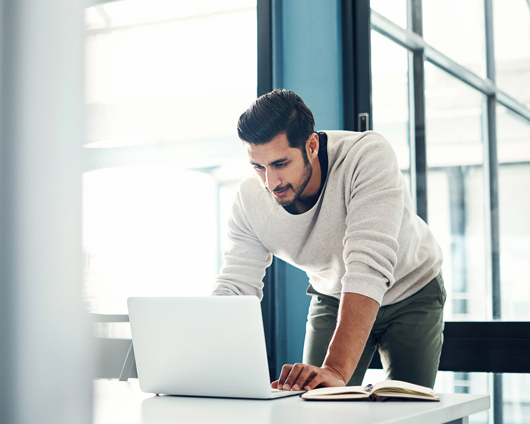 Cloud – Against a backdrop of windows, a man bends forward to lean against a desk as he looks at his laptop