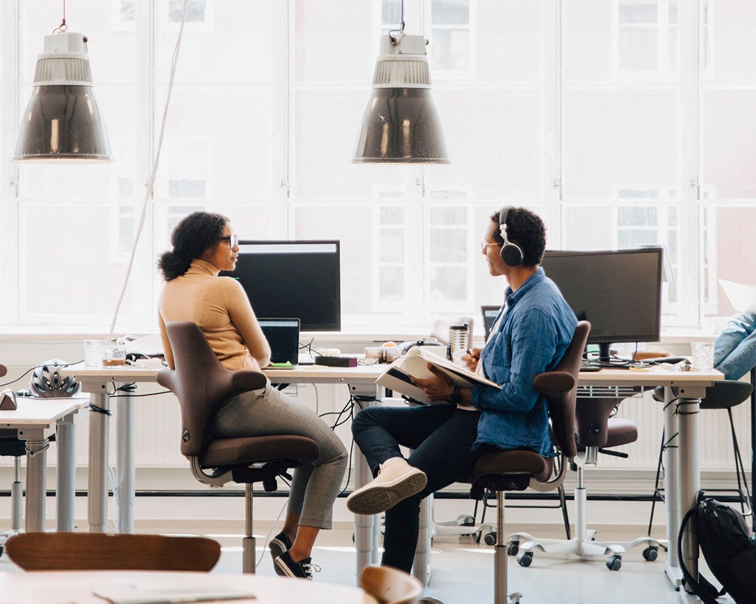 Cloud adoption – Two colleagues sit next to each other at a workdesk facing a bright window