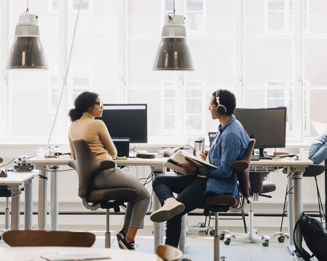 Cloud adoption – Two colleagues sit next to each other at a workdesk facing a bright window