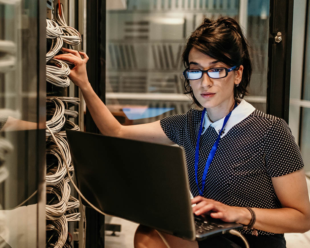 Database Security – Woman in glasses typing on laptop in server room.