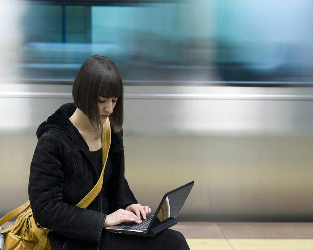 Fast Recovery – A woman sits on a bench working on a PC laptop as a subway train speeds behind her