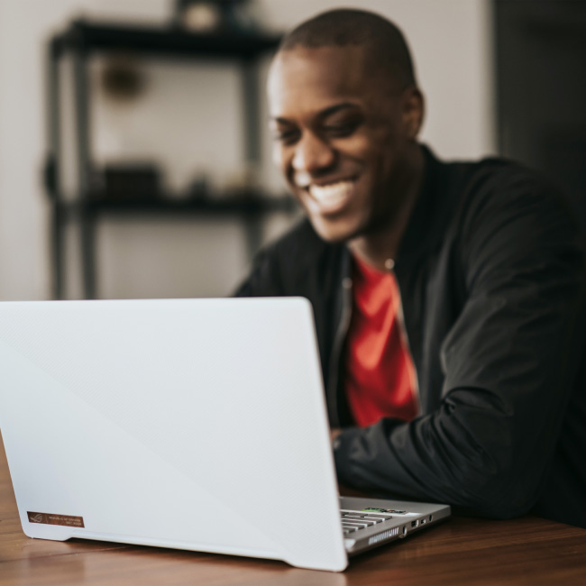 A man with a cheerful expression sits at a table, engrossed in his work on a laptop.