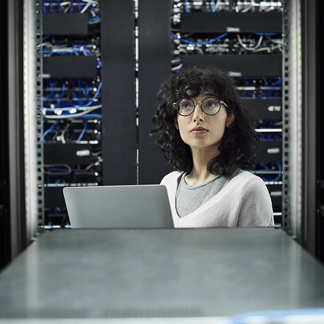 A woman standing in a  server room in front of some blue cables, holding a laptop computer