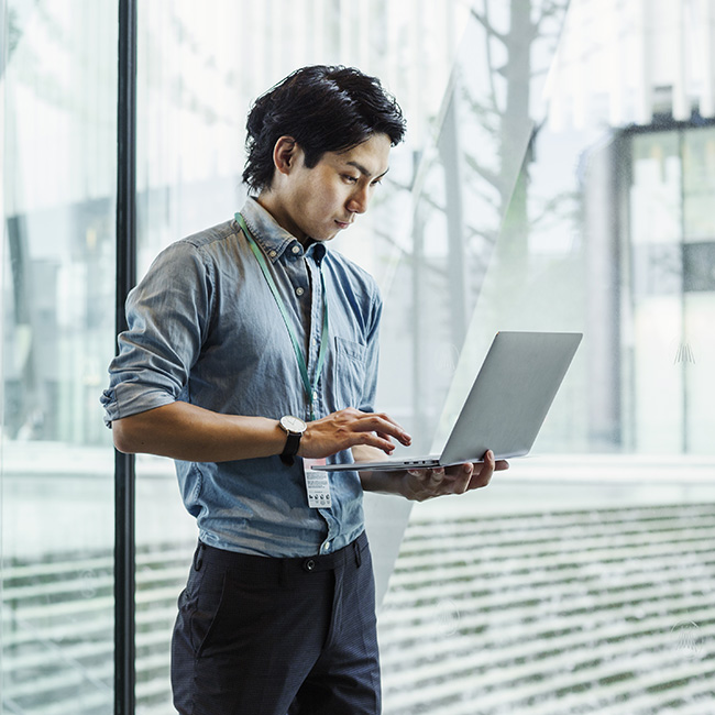 A man works on a laptop, standing in front of a glass window in an ultramodern building