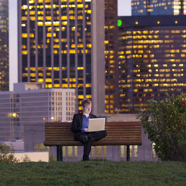 A man sits on a bench in a park in the middle of an urban area, working on a laptop