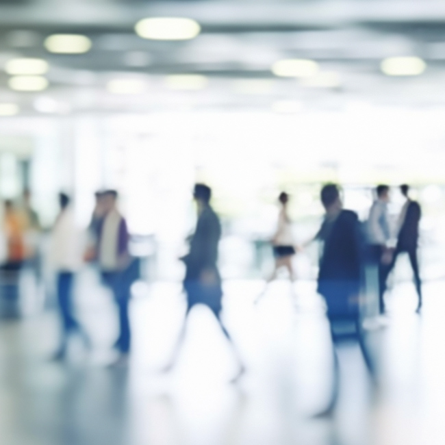 Many blurred people walk through a bright lobby area in an office building with a bustling work environment