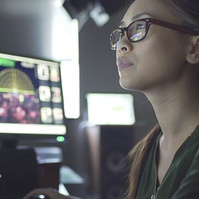 A smart woman wearing glasses confidently gazes at a set of computer screens