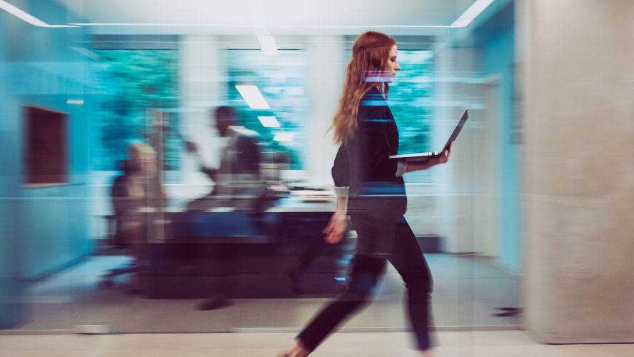 A woman confidently walks through an office building, carrying a laptop, engrossed in her work.