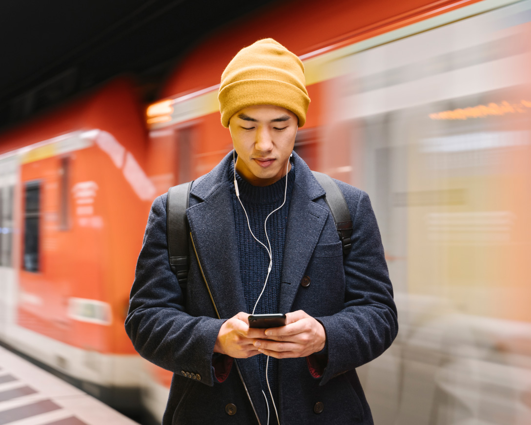 A man wearing a beanie and jacket is seen using his phone in front of a train.