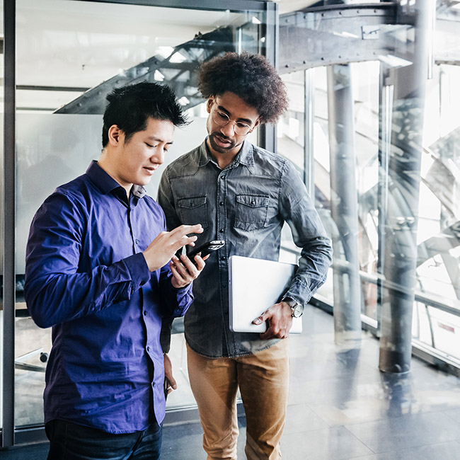 A man holding a laptop stands next to a colleague as they review the contents of a smartphone