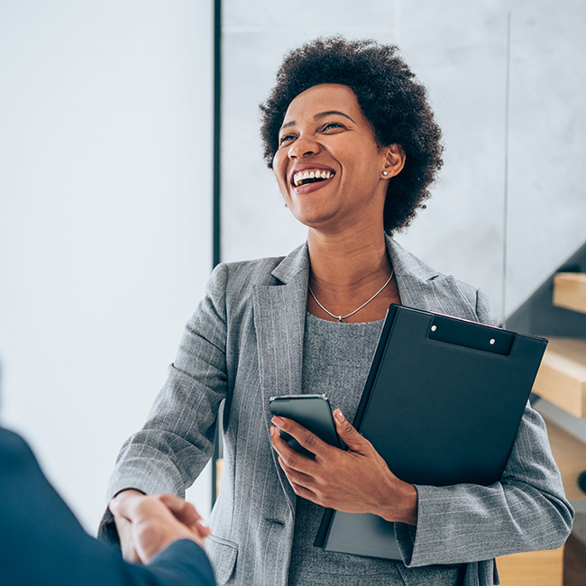 A smiling woman holding a notepad and phone shakes the had of a business partner