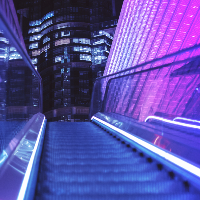 Image depicts a view of a brightly-lit escalator well looking up towards the city skyline.