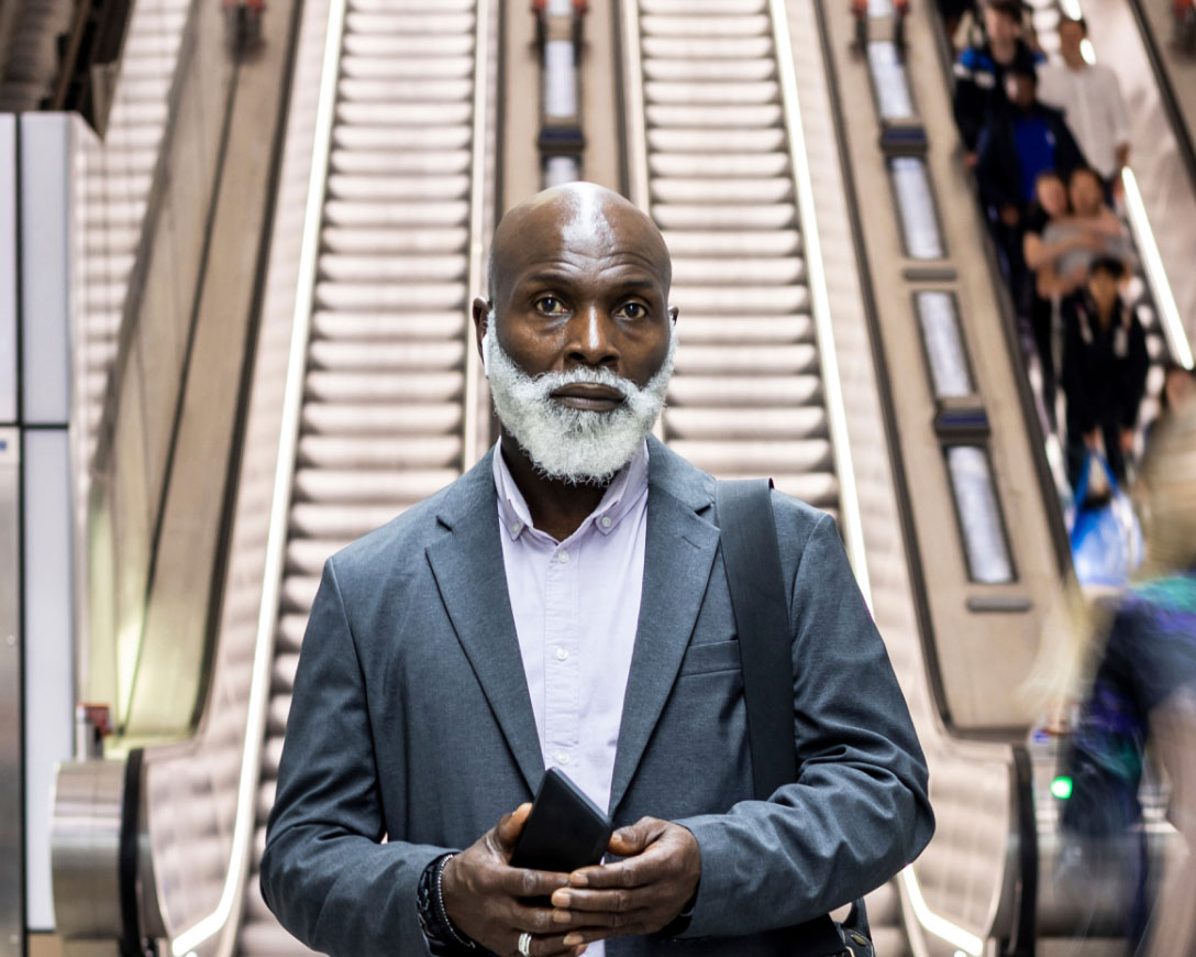 Grey-bearded man standing in front of an escalator.