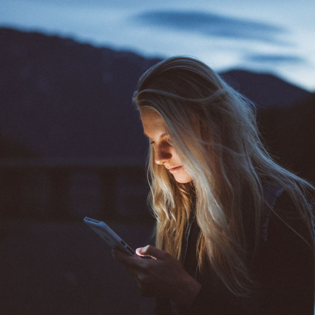 A woman engrossed in her phone's screen, captivated by its glow, as darkness surrounds her during the night.