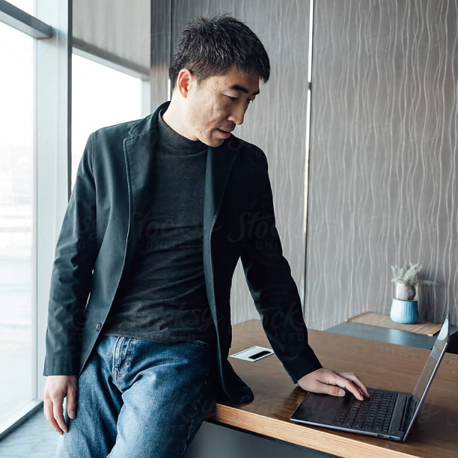A man in casual attire, wearing jeans and a jacket, is seated at a desk, engrossed in his work on a laptop.