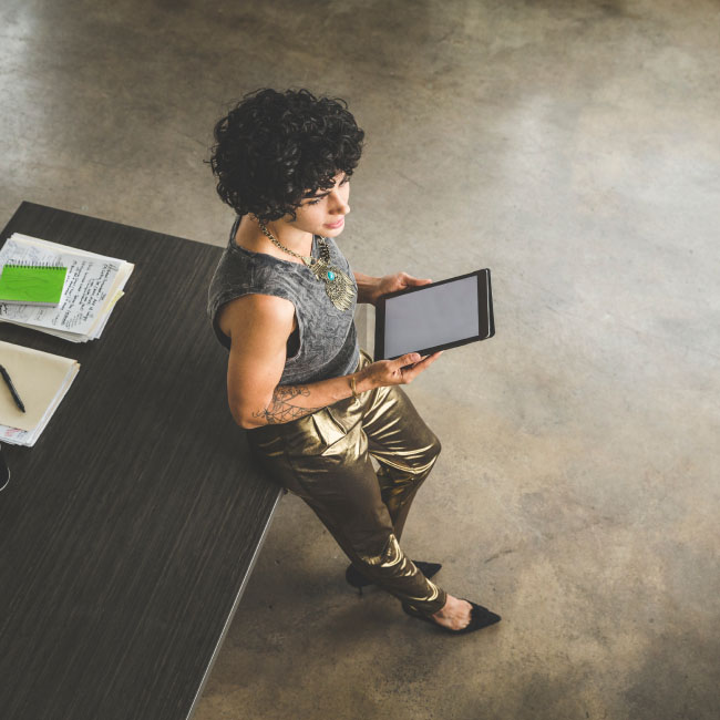 A woman sitting on a desk, engrossed with a tablet, displaying focus and productivity.